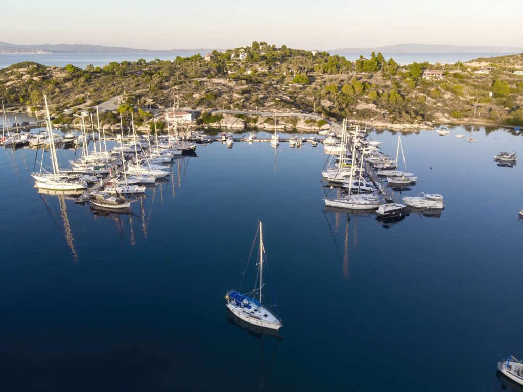 aegean-sea-port-with-multiple-moored-yachts-near-piers-greenery-blue-water-view-from-drone-greece